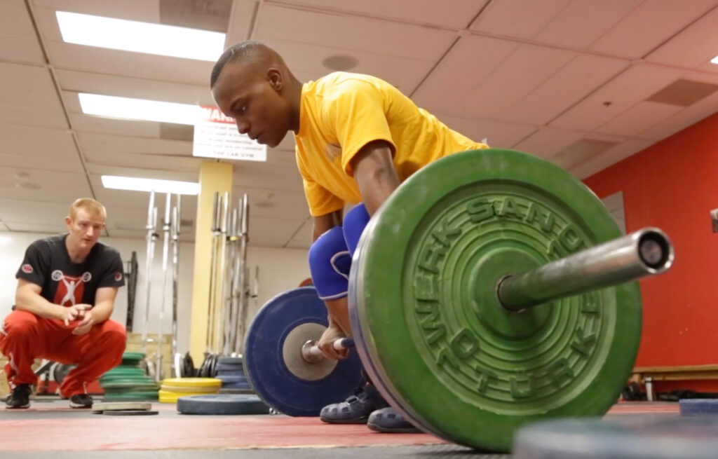 Jerome about to perform a deadlift with large weights and coach observing nearby